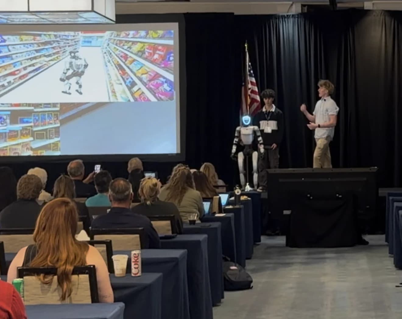 Presenters demonstrating Project Meeseeks on stage, with retail testing footage on the screen and an audience in front.